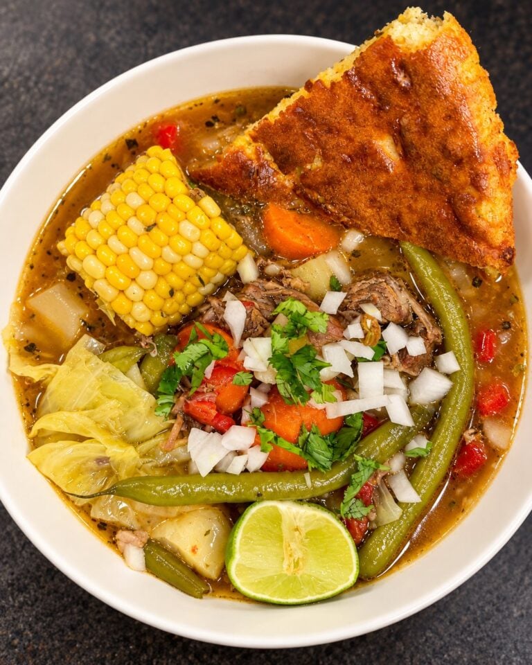 Bowl of caldo de res with beef, vegetables, fresh garnishes, lime, and jalapeño cornbread